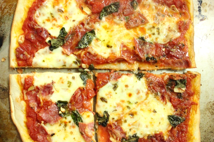 An overhead of a simple red-sauce pizza with basil and thin-sliced Italian meats.